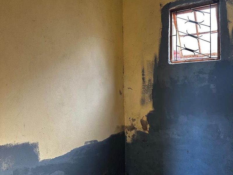 An empty room featuring unfinished wall painting, exposed concrete flooring, and a window with security bars, requiring significant renovation and cosmetic updates.