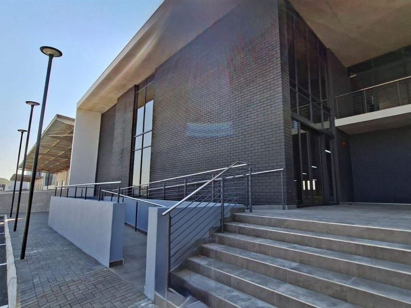 Modern industrial building exterior featuring a dark brick facade, concrete staircase, accessible ramp with metal railings, and paved parking area.