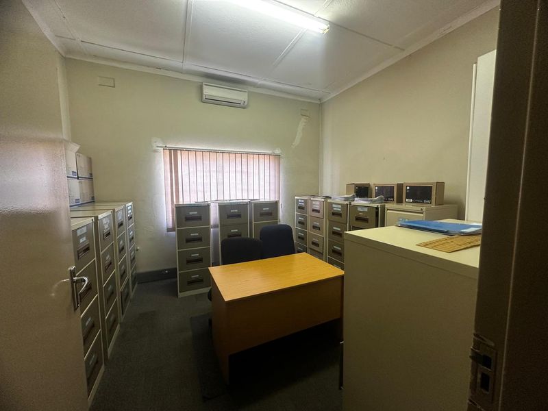 A functional office space featuring carpeted flooring, a wall-mounted air conditioning unit, and vertical blinds, currently utilized for storage and administrative tasks.