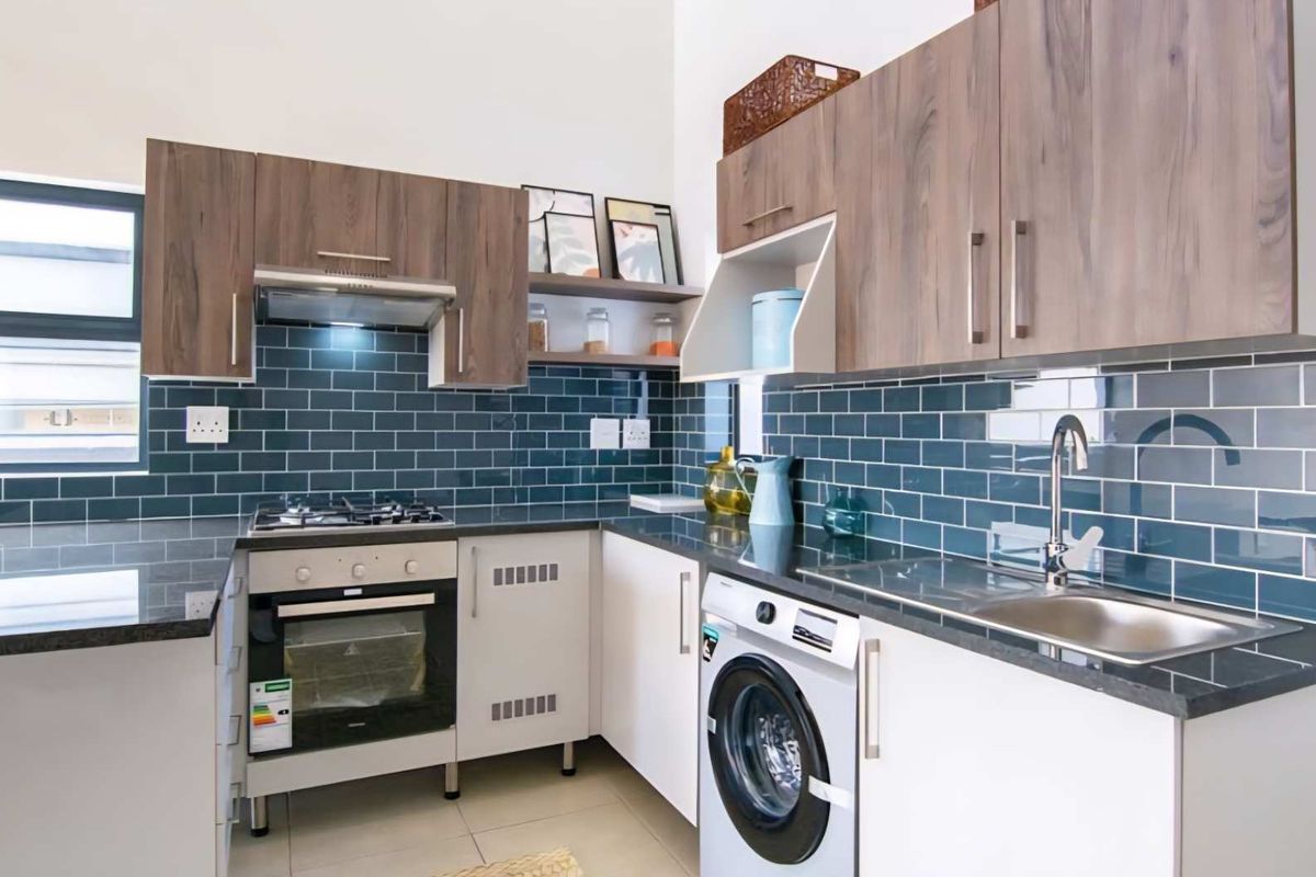 Modern kitchen featuring dark stone countertops, blue subway tile backsplash, built-in oven with gas hob, extractor fan, and integrated laundry appliance space.