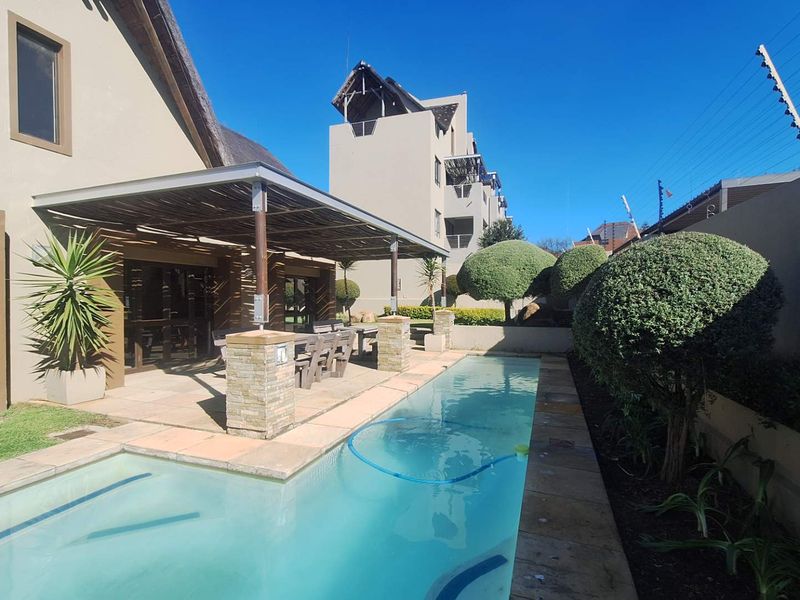 Outdoor pool area featuring a swimming pool, covered patio with stone-clad pillars, and landscaped garden borders within a secure complex.