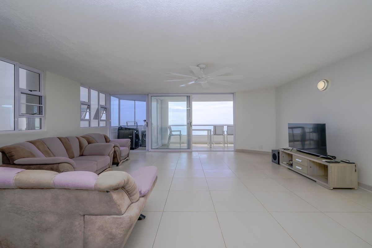 Spacious open-plan living area featuring neutral tiled flooring, a ceiling fan, and large sliding glass doors leading to a balcony with ocean views.