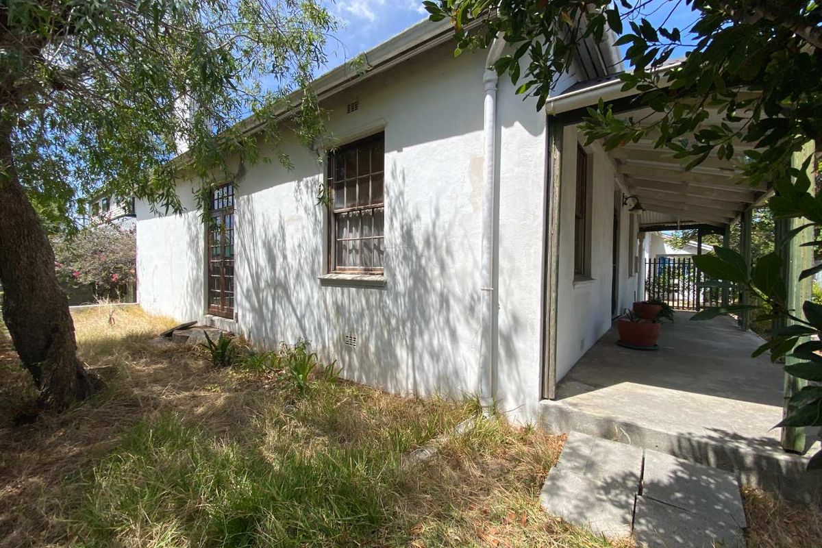 Exterior view of the house featuring a covered patio area, white rendered walls, and surrounding garden space requiring maintenance.