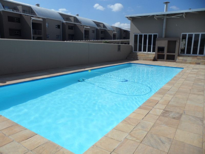 A well-maintained swimming pool area featuring a paved deck, built-in braai, and secure boundary wall within a residential apartment complex.