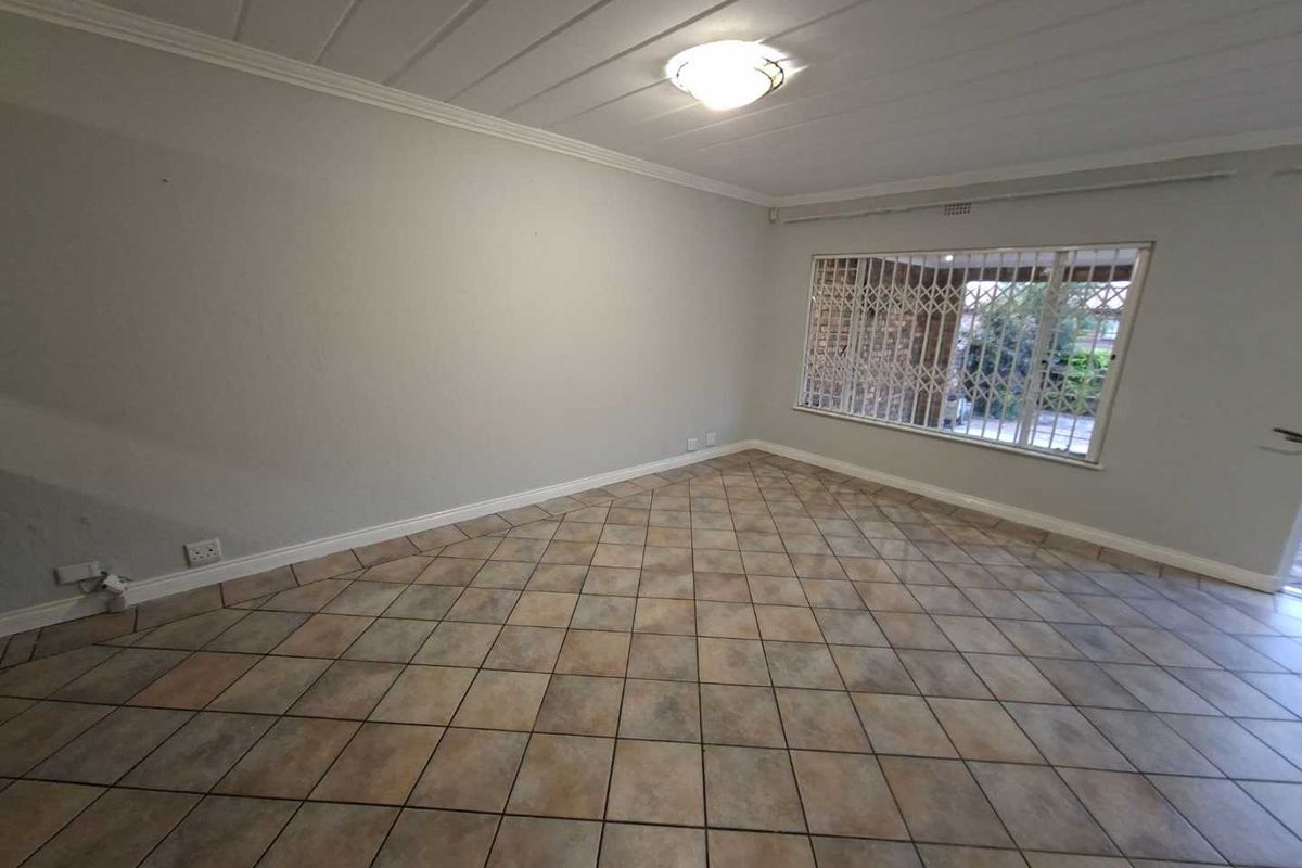 A spacious, empty living room featuring neutral tiled flooring, coved ceilings, and large windows fitted with security bars.