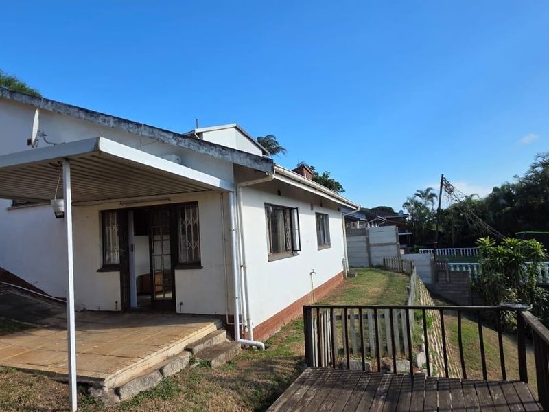 Rear view of the property featuring a covered patio area, timber decking, and a garden space with retaining walls.