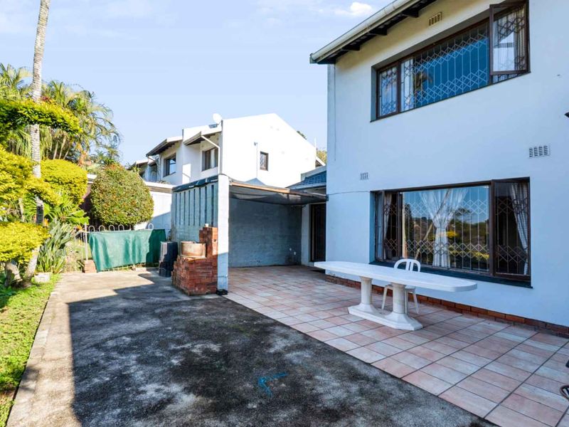 Rear exterior view of the townhouse featuring a tiled patio, a brick built-in braai area, and a concrete paved section leading to the garden.