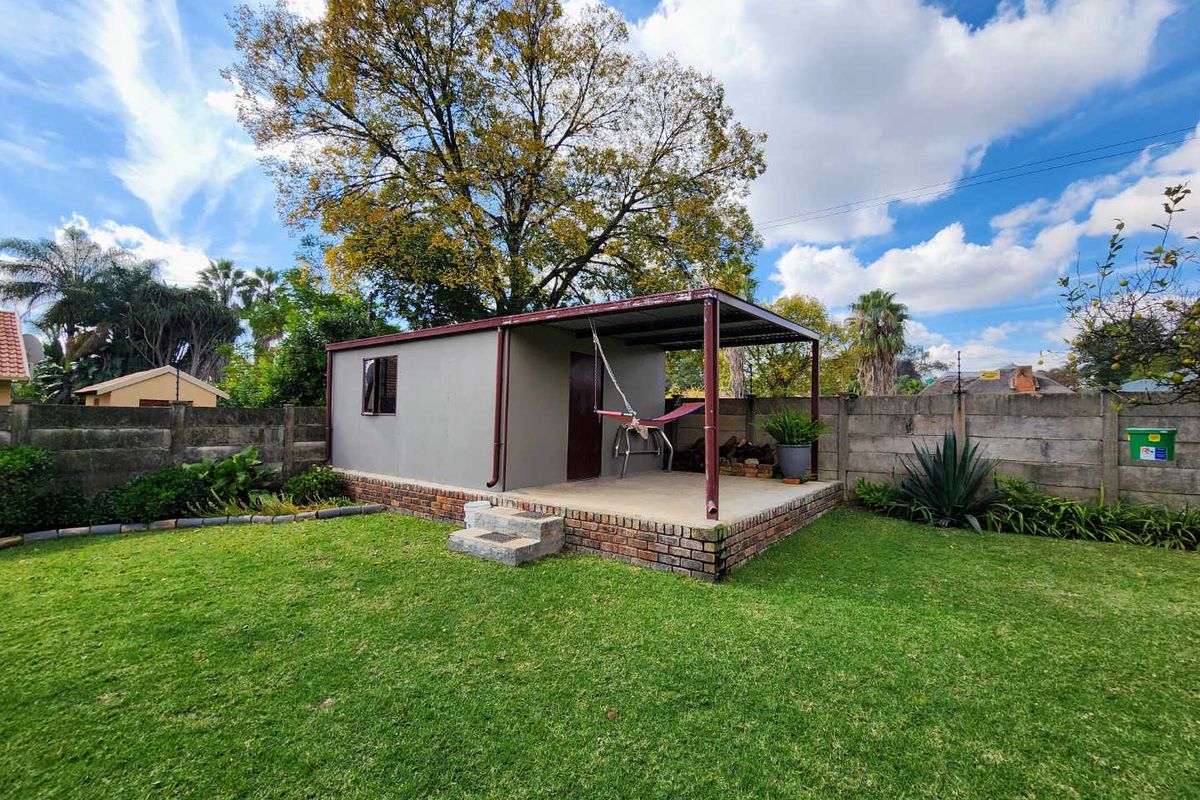 A detached garden outbuilding featuring a covered patio area, brick foundation, and concrete flooring, set within a well-maintained lawn.
