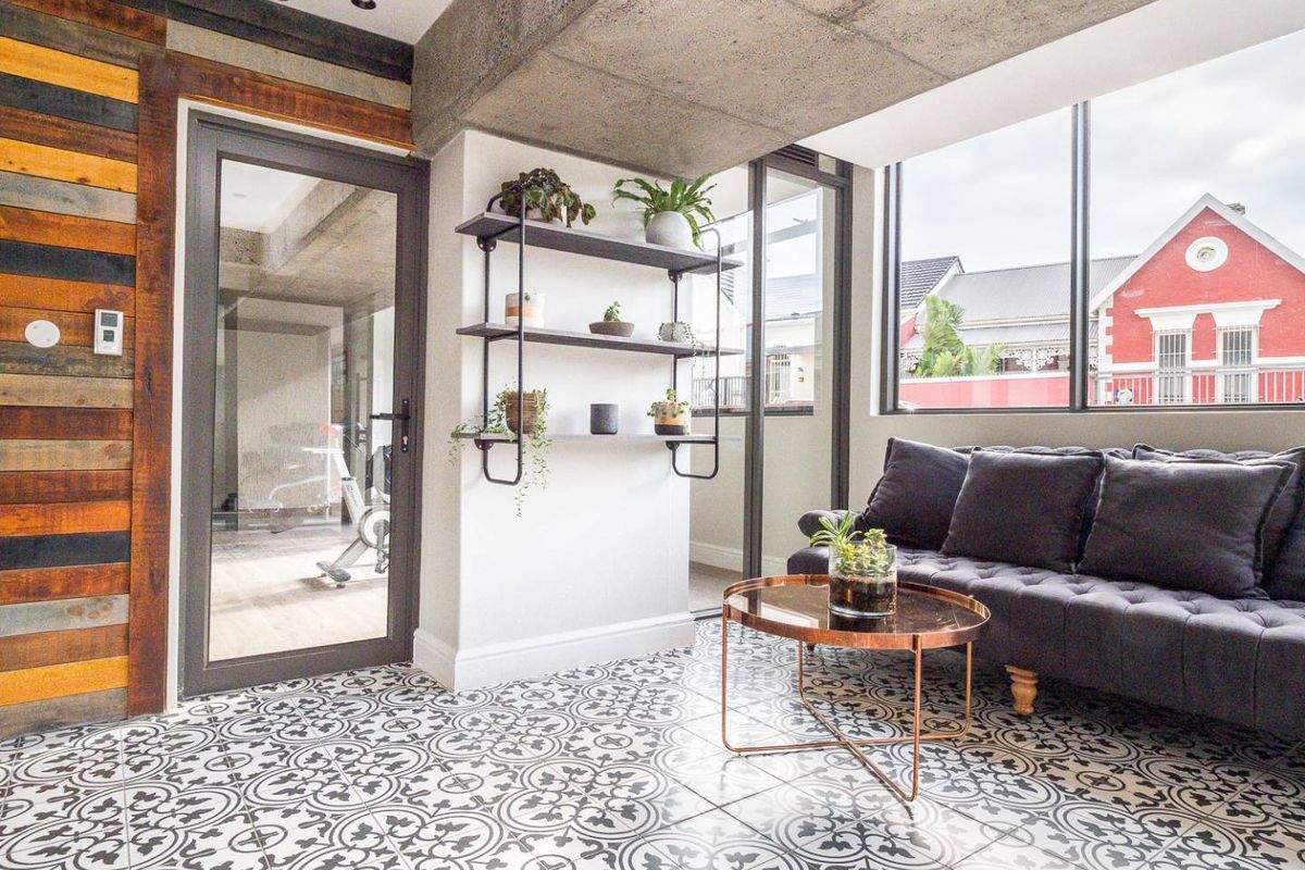 A modern living area featuring decorative patterned floor tiles, exposed concrete ceiling beams, and a glass door leading to an adjacent room.