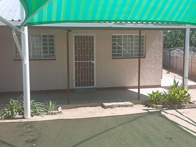 Covered patio area featuring a green shade sail, security gate, and burglar bars, overlooking a simple backyard with perimeter walling.