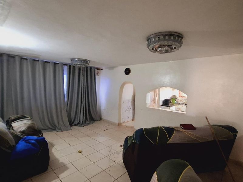 Living room featuring tiled flooring, an arched doorway, and a pass-through window to the kitchen area, requiring significant cosmetic updates and repairs.