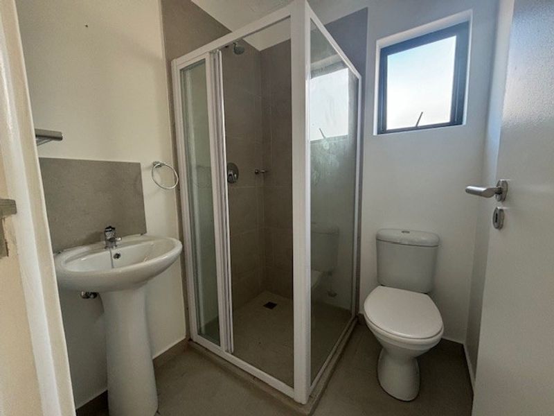 A functional bathroom featuring a pedestal washbasin, a glass-enclosed shower, and a standard toilet with neutral tiled finishes.