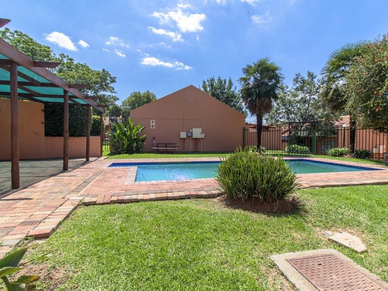 A communal outdoor pool area featuring a swimming pool, brick paving, and a wooden pergola set within a landscaped garden.