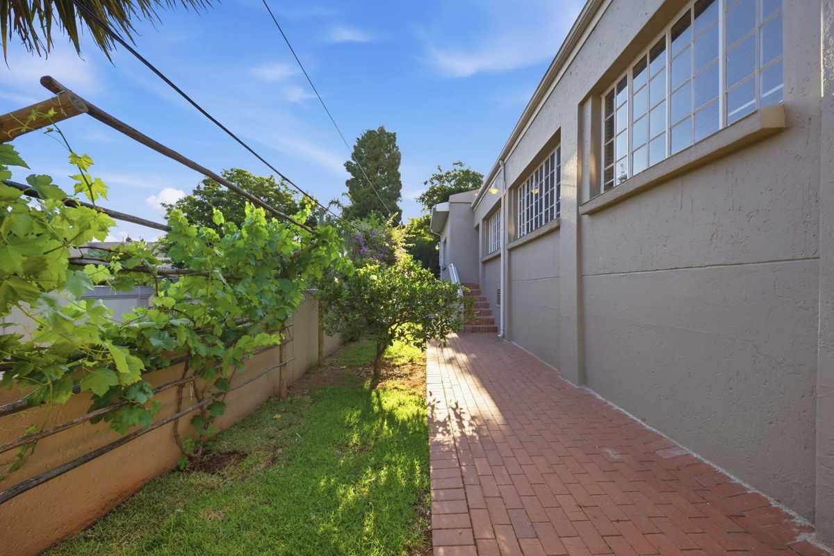 A well-maintained rear exterior view featuring a brick paved walkway, boundary wall with trellis, and multi-pane windows along the side of the house.