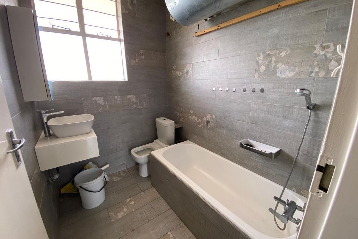 A compact bathroom featuring grey wood-look wall and floor tiles, a white bathtub with shower attachment, a wall-mounted basin, and a toilet.