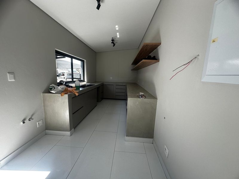 A modern, minimalist scullery featuring concrete-look cabinetry, floating timber shelving, tiled flooring, and an undermount sink, currently awaiting final electrical finishing.