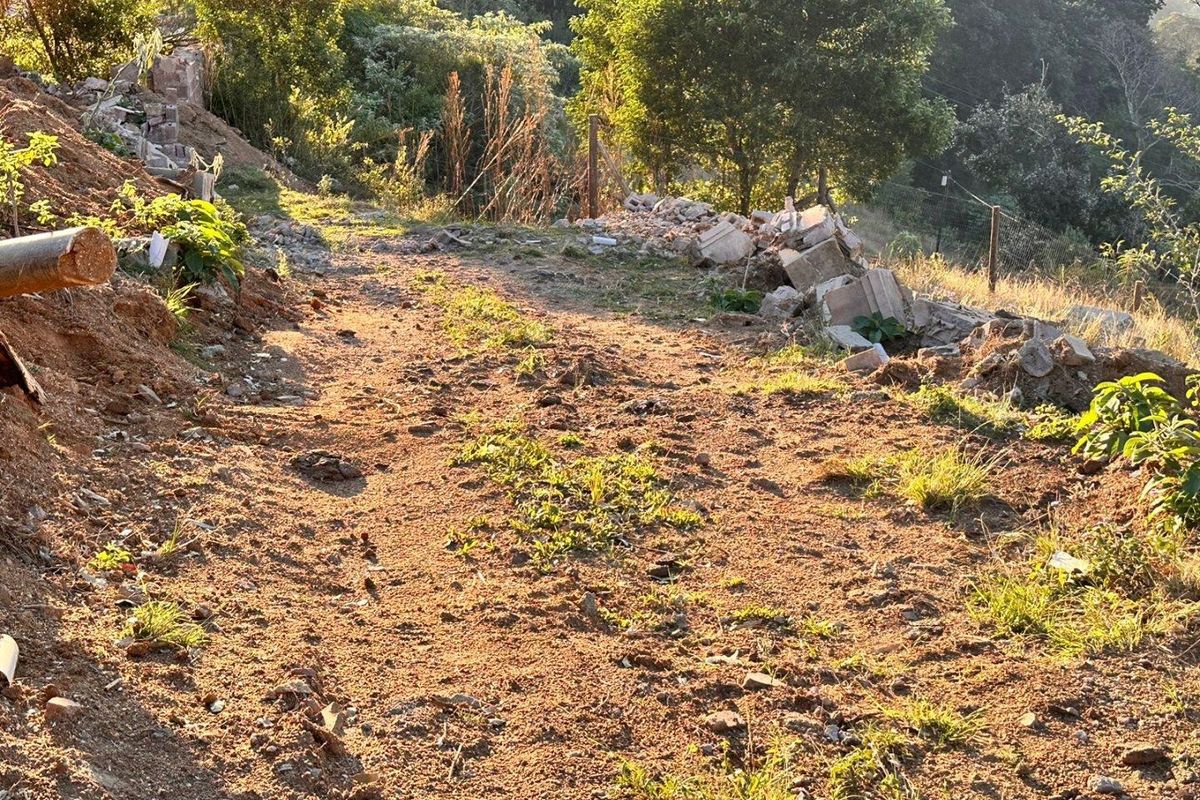 A view of a sloped residential plot featuring natural terrain, scattered debris, and existing boundary fencing.