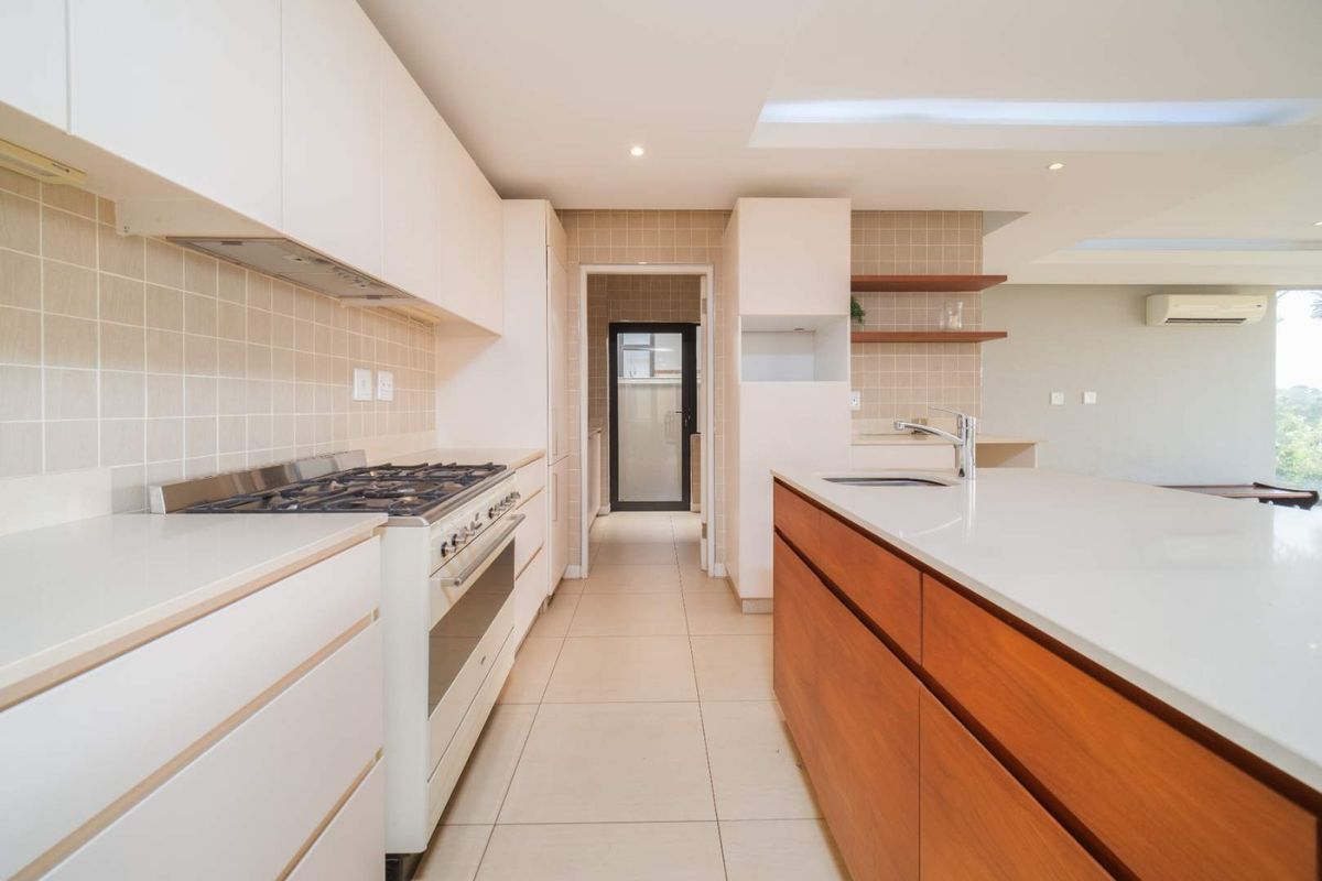 Modern kitchen featuring a central island with stone countertops, gas stove, tiled backsplash, and sleek cabinetry, leading to a scullery area.