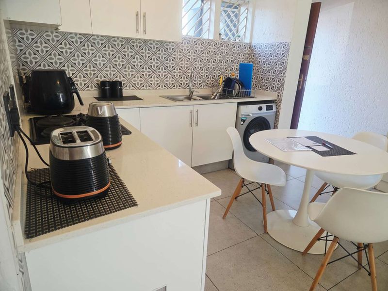 A functional kitchen featuring white cabinetry, stone countertops, a patterned tile backsplash, and an integrated washing machine, with an adjacent dining area.