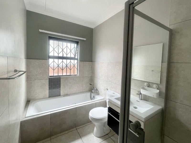 A clean and functional bathroom featuring a built-in bathtub, vanity basin, toilet, and neutral tiled finishes with security bars on the window.