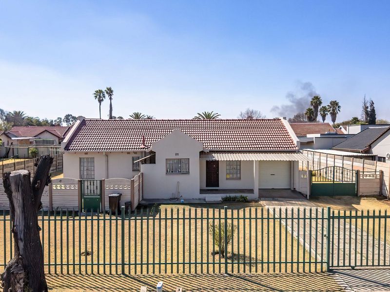 Front view of a single-story residential house featuring a tiled roof, attached garage, paved driveway, and secure perimeter fencing.