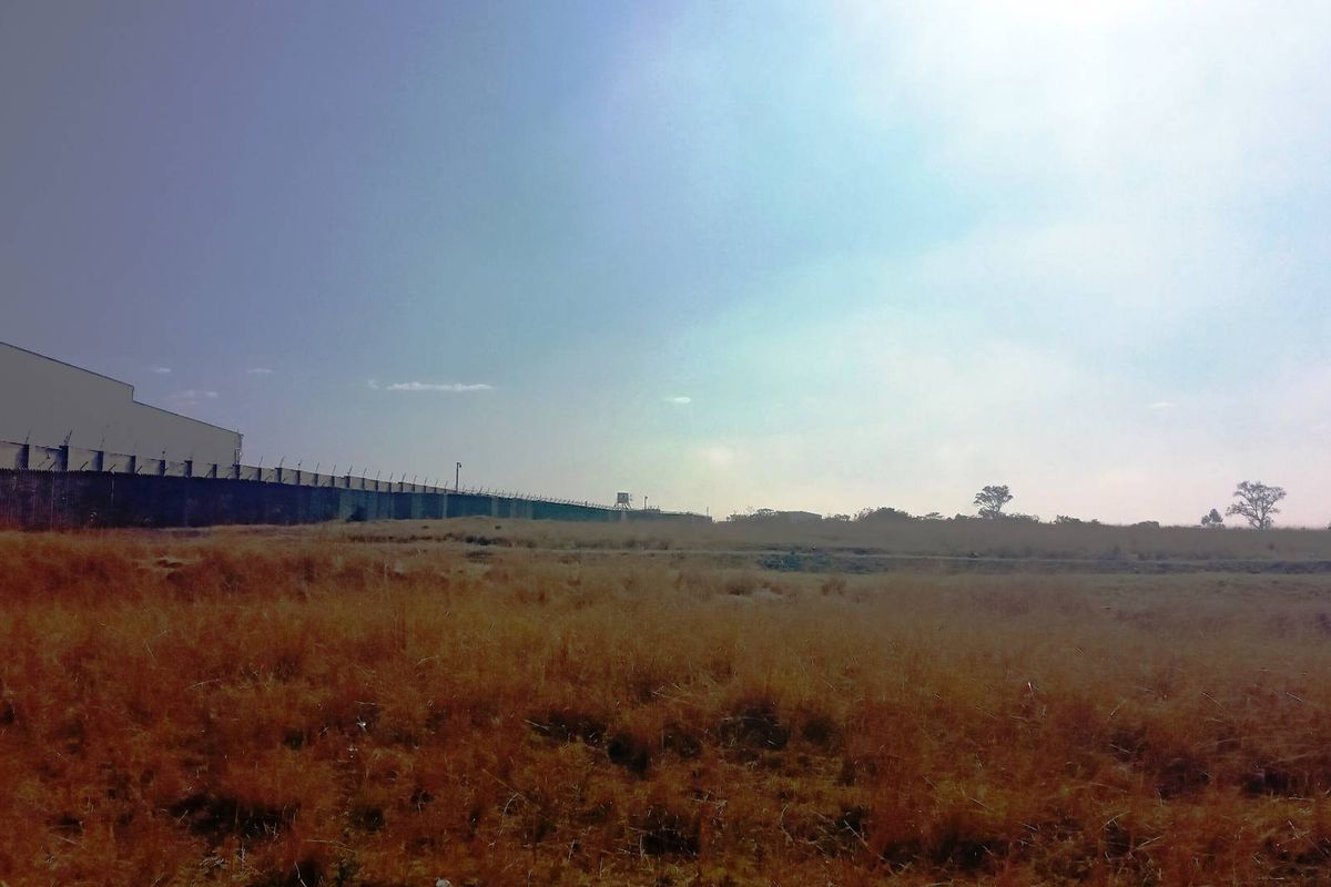 A wide view of a vacant commercial land plot featuring dry grass and perimeter security fencing along the boundary.