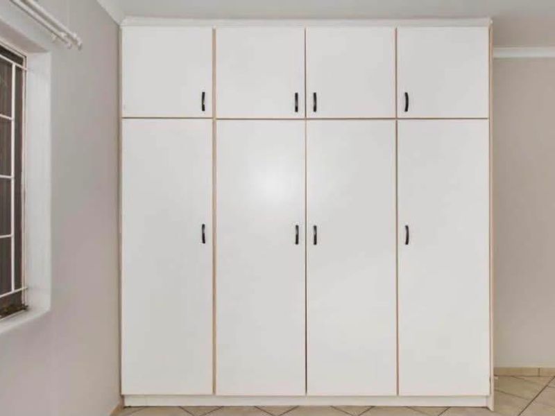 A clean bedroom featuring floor-to-ceiling built-in cupboards, neutral tiled flooring, and a window fitted with security bars.