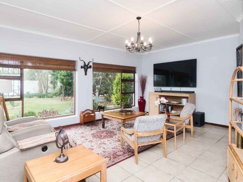 A bright and spacious living room featuring large windows with garden views, tiled flooring, and a classic chandelier.