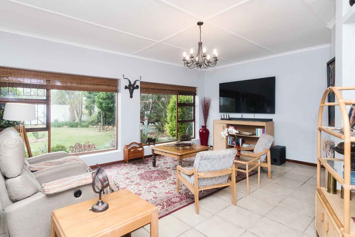 A bright and spacious living room featuring large windows with garden views, tiled flooring, and a classic chandelier.