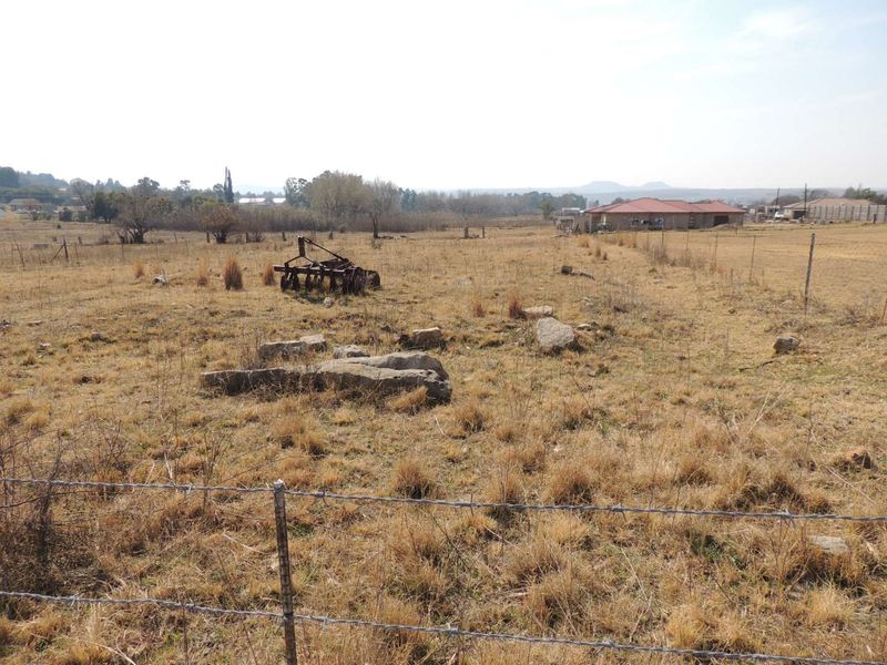 A view of a vacant residential plot featuring dry grass, scattered rocks, and perimeter wire fencing with neighboring properties visible in the distance.