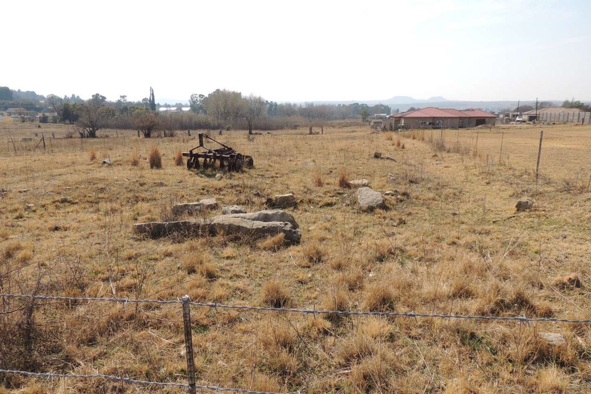 A view of a vacant residential plot featuring dry grass, scattered rocks, and perimeter wire fencing with neighboring properties visible in the distance.