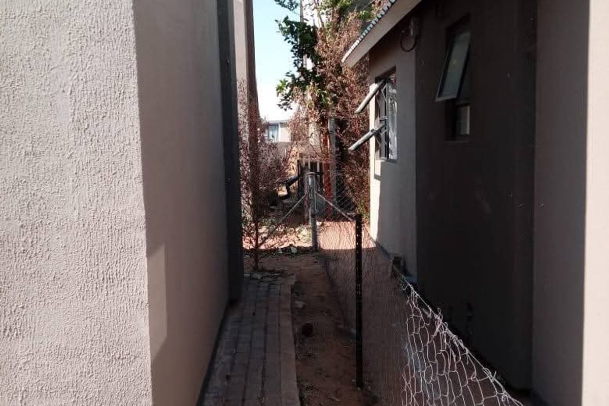 A narrow side passage between townhouse units featuring a paved walkway and wire mesh boundary fencing.