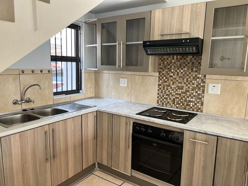 A functional kitchen featuring wood-look cabinetry, mosaic tile backsplash, built-in oven with hob, and a double stainless steel sink under a window with security bars.