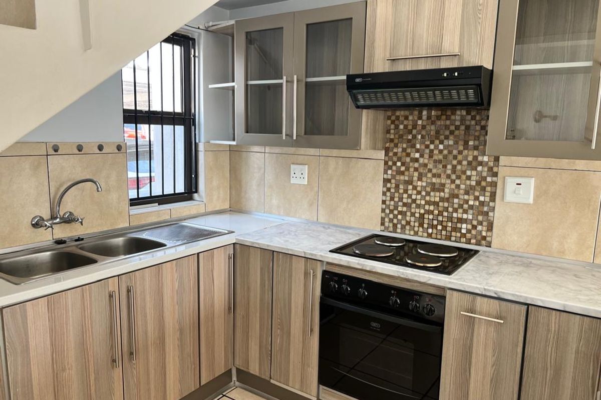 A functional kitchen featuring wood-look cabinetry, mosaic tile backsplash, built-in oven with hob, and a double stainless steel sink under a window with security bars.