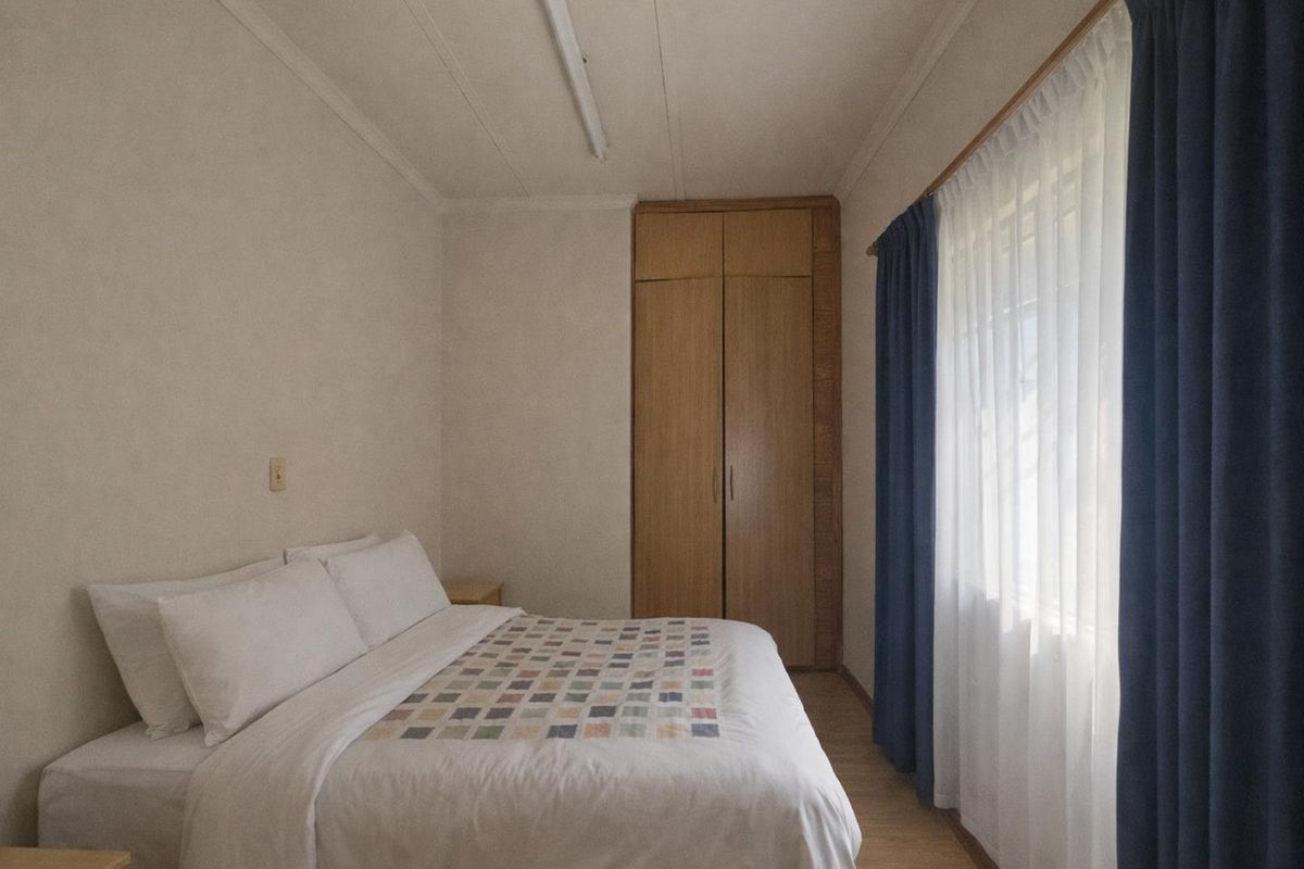 A functional bedroom featuring built-in wooden cabinetry, laminate flooring, and large windows with curtains, offering a simple and practical living space.