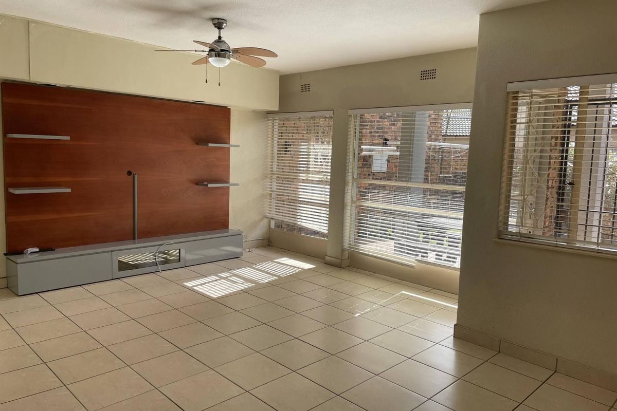 Spacious living area featuring neutral floor tiles, a built-in wooden media wall unit, ceiling fan, and large windows with fitted blinds.