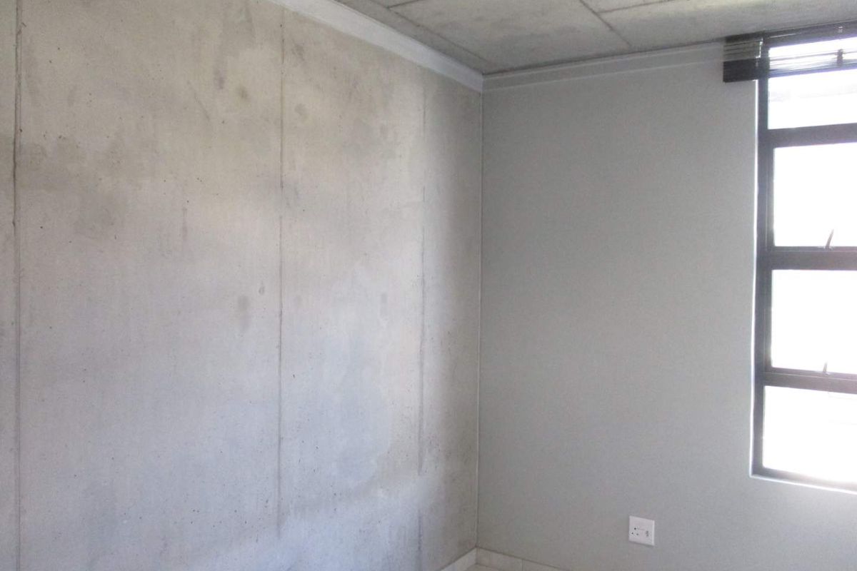 A minimalist bedroom featuring an exposed concrete feature wall, tiled flooring, and a large window providing natural light.