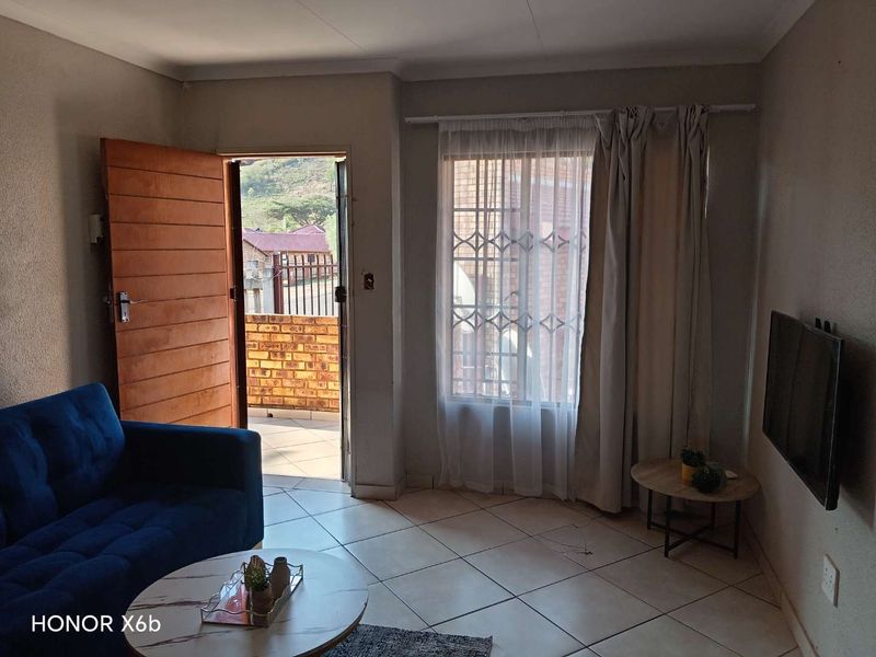 A living room featuring tiled flooring, a timber entrance door, and a window with security burglar bars, providing access to an outdoor area.