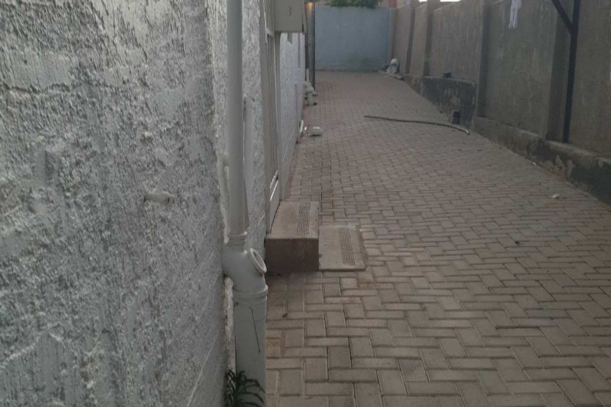 A narrow side exterior walkway featuring paved ground and a textured exterior wall with exposed plumbing fixtures.
