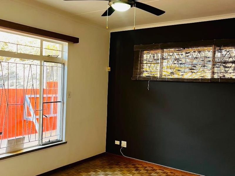 A bright bedroom featuring polished parquet flooring, a ceiling fan, and a dark accent wall with security bars on the window.