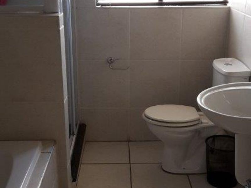 A functional bathroom featuring a shower enclosure, pedestal basin, and toilet, finished with neutral wall and floor tiles and fitted with venetian blinds.