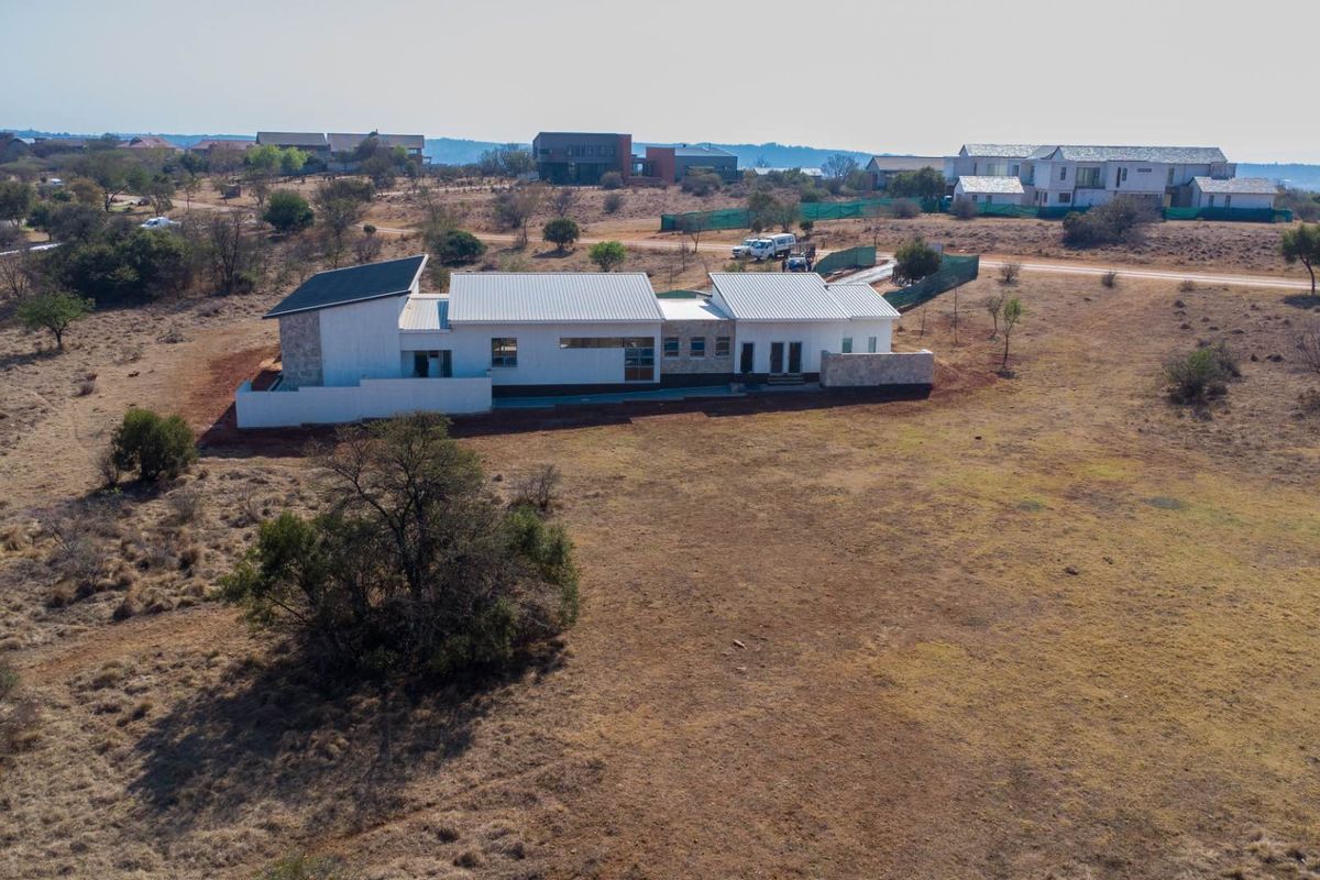 Aerial view of a modern residential property featuring a metal roof, stone cladding, and white exterior walls situated on a large vacant plot.