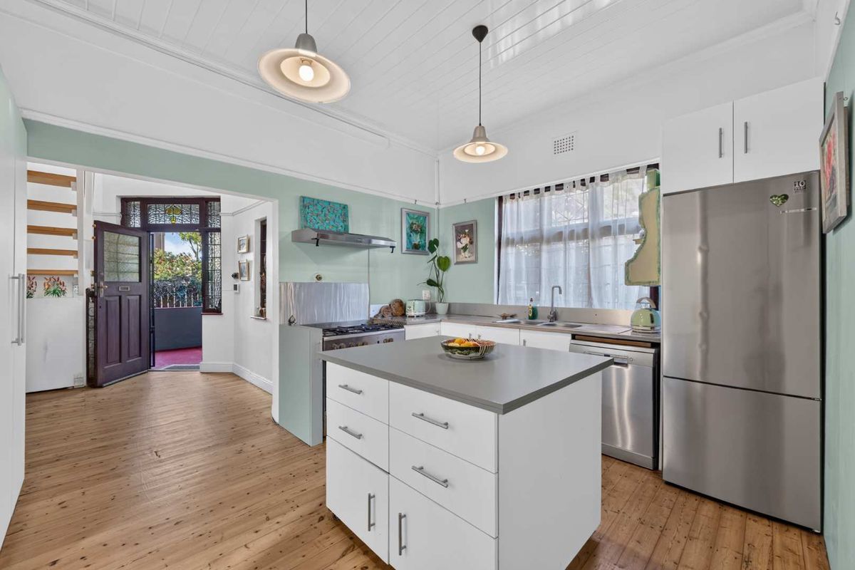 A bright kitchen featuring a central island, stainless steel appliances, timber flooring, and classic tongue and groove ceiling details, with direct access to the entryway.