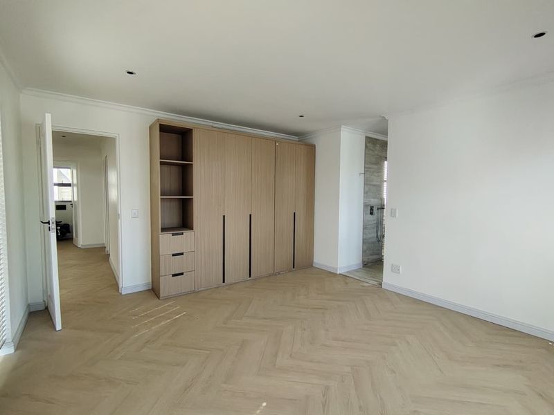 A bright, modern bedroom featuring light wood-tone herringbone flooring, built-in cabinetry, and clean white walls, leading into an adjacent en-suite bathroom area.