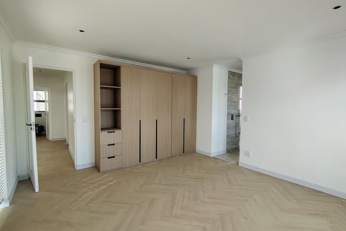 A bright, modern bedroom featuring light wood-tone herringbone flooring, built-in cabinetry, and clean white walls, leading into an adjacent en-suite bathroom area.