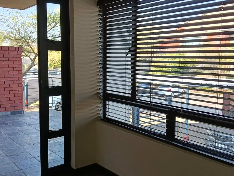 A covered balcony area featuring tiled flooring, a brick feature wall, and large windows with fitted venetian blinds, offering a bright and functional outdoor space.