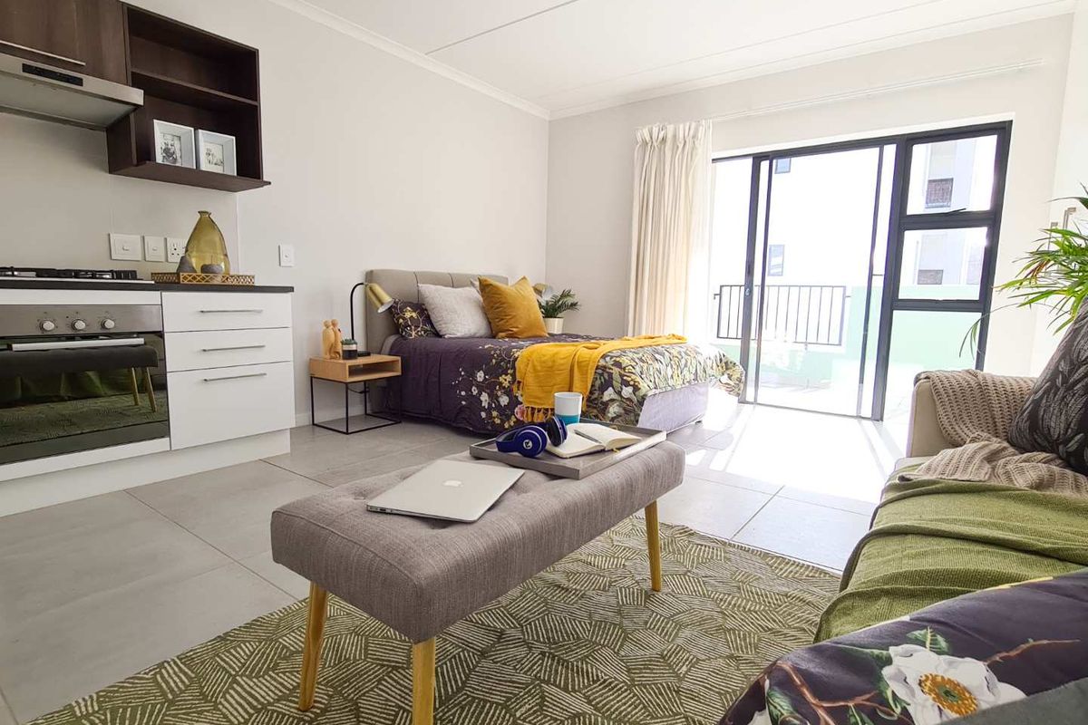 Modern open-plan studio apartment featuring a compact kitchen with built-in appliances, tiled flooring, and direct access to a private balcony through sliding glass doors.