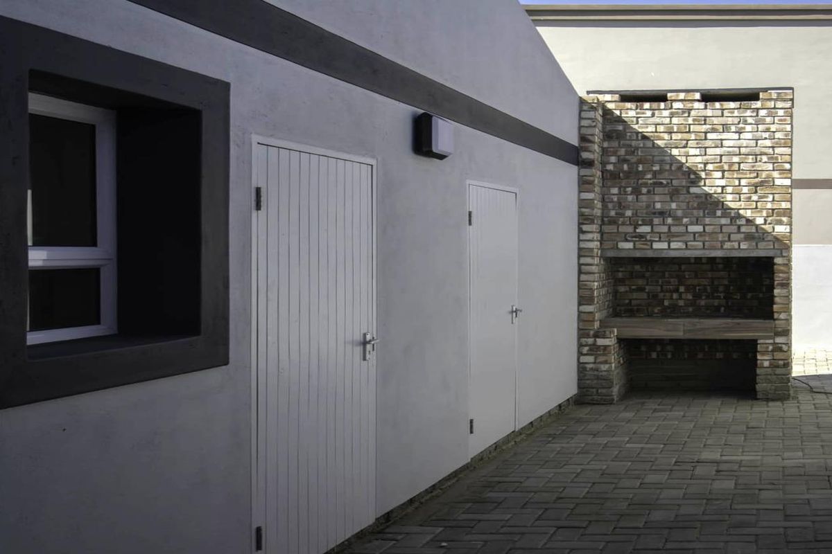 A paved outdoor courtyard area featuring a built-in brick braai, exterior wall lighting, and access doors to the property.