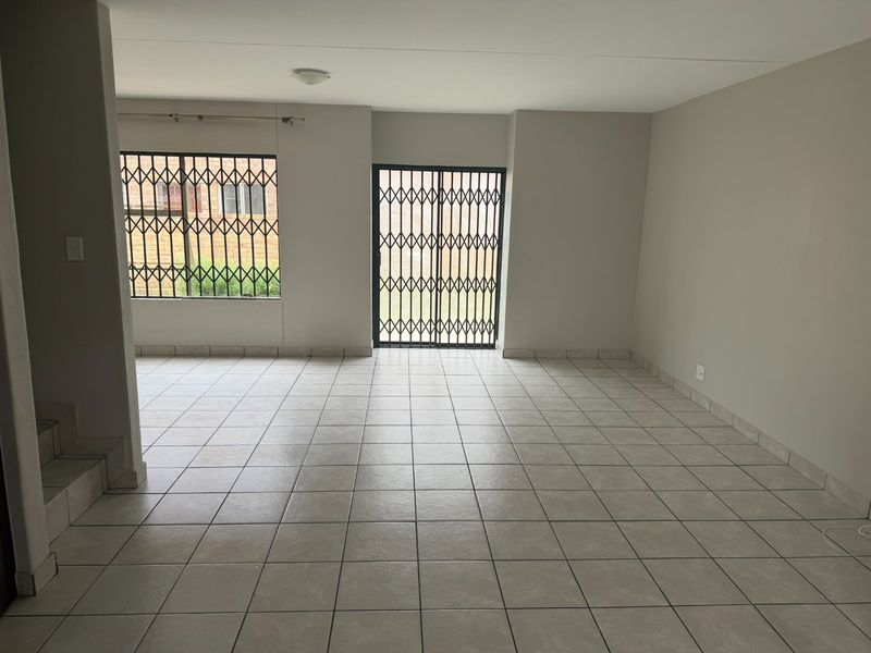 An empty, open-plan living area featuring neutral ceramic floor tiles, security gates on the sliding door, and security bars on the windows.