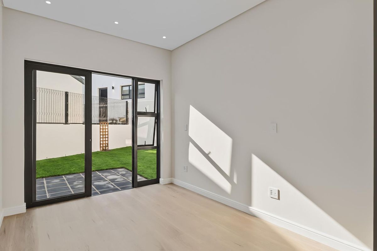 A bright, empty living room featuring light wood-look flooring, recessed ceiling lighting, and large sliding glass doors opening onto a private garden patio.